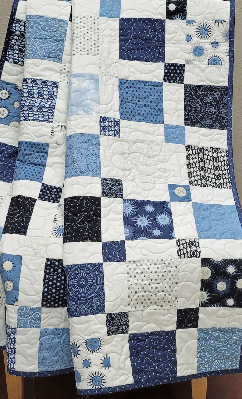 May include: A patchwork quilt in shades of blue and white, featuring various celestial patterns. The quilt is composed of square blocks with designs of suns, moons, stars, and zodiac symbols. The quilt is quilted with a wavy pattern.
