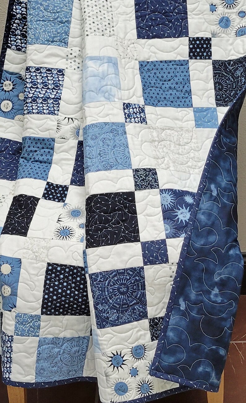 May include: A patchwork quilt in shades of blue and white, featuring celestial and geometric patterns. The quilt's design includes squares with moons, stars, and zodiac symbols. The edges are finished with a dark blue fabric with a cloud-like pattern.