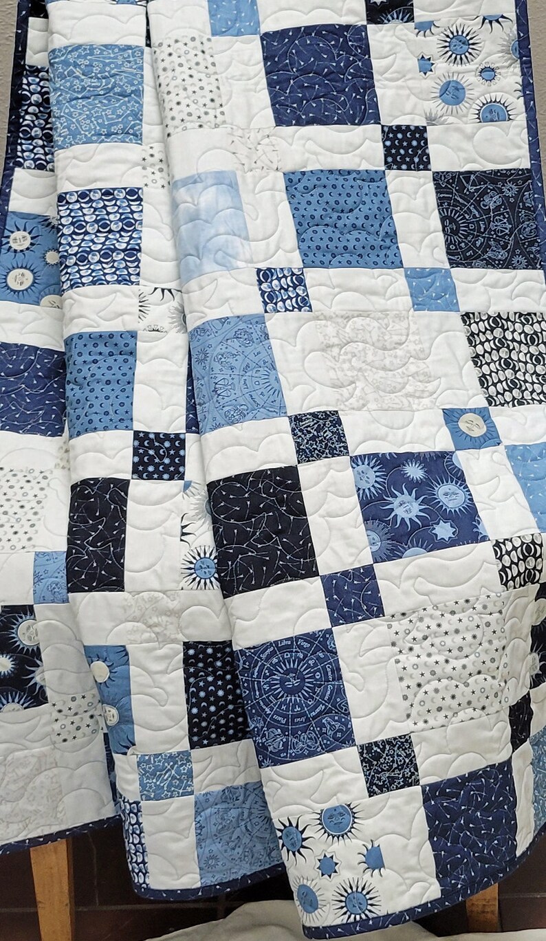 May include: A patchwork quilt featuring squares in shades of blue and white. The squares display celestial patterns like suns, moons, stars, and zodiac symbols. The quilt is quilted with a wavy pattern.