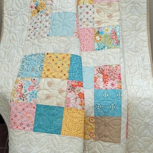 May include: A colourful patchwork quilt with a cream background and various patterned squares. The squares incorporate floral, striped, and patterned fabrics in pink, blue, yellow, and brown. The quilt has a scalloped edge.