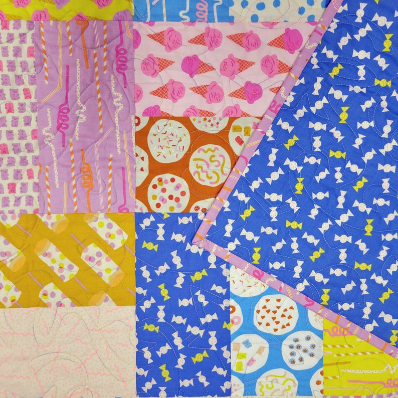 Pu&ograve; includere: Una colorata trapunta patchwork con vari motivi, tra cui coni gelato, caramelle e cannucce. La trapunta presenta un motivo di caramelle blu sull'angolo, con un bordo rosa. Il design incorpora tonalit&agrave; rosa, arancioni, gialle e viola.