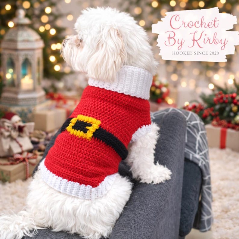 May include: A small white dog wearing a red and white crocheted Santa Claus outfit with a black belt and yellow buckle. The text "Crochet By Kirby" and "Hooked Since 2020" are visible. The background has Christmas lights and wrapped gifts.