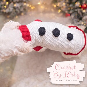 May include: A white crochet dog sweater with three black dots and red trim. The sweater is designed to look like a snowman. The text "Crochet By Kirby" is visible in the lower right corner. The sweater is designed for a small dog.