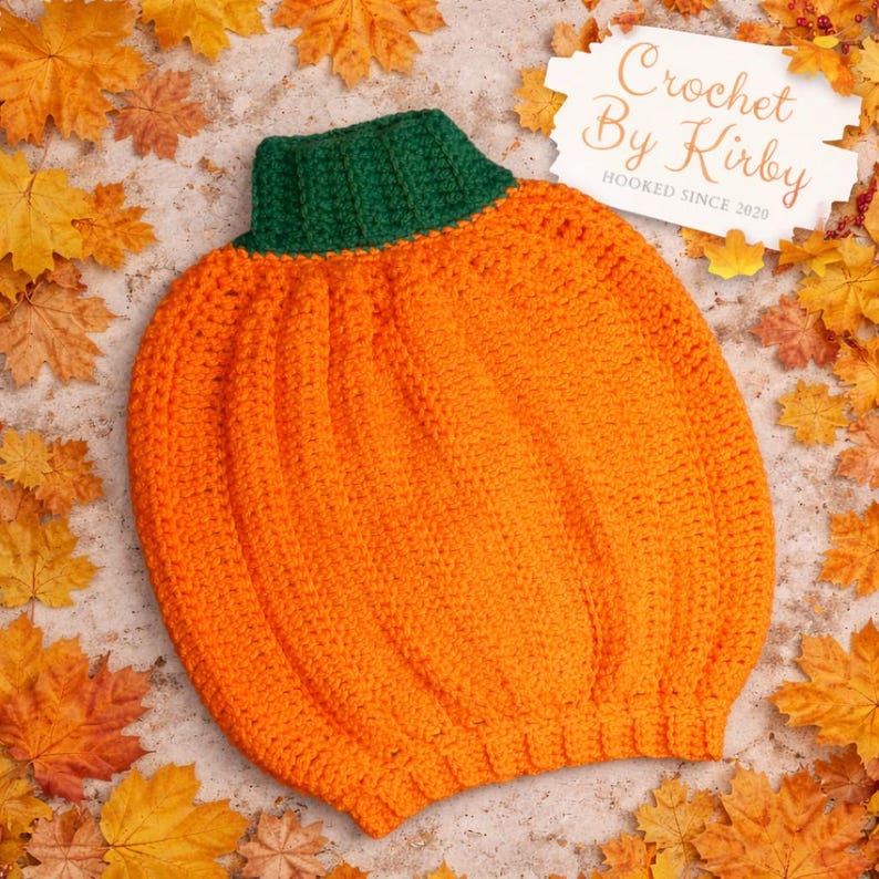 May include: An orange crochet pumpkin hat with a green collar. The hat is surrounded by autumn leaves. The text "Crochet By Kirby Hooked Since 2020" is in the upper right corner.