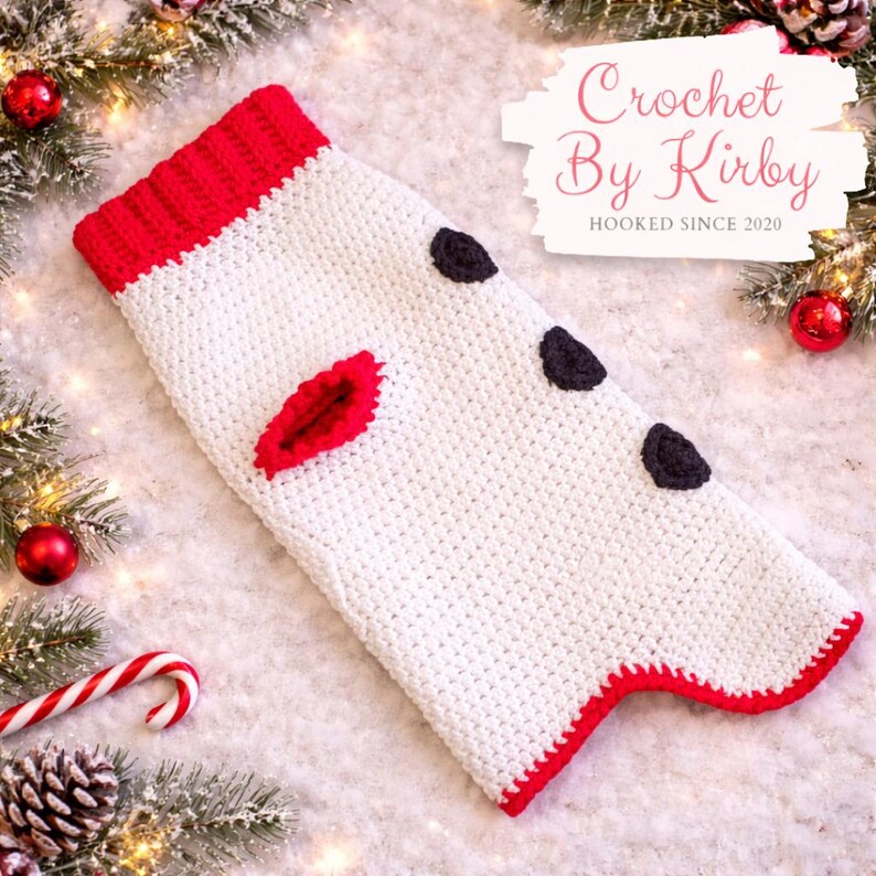 May include: A handmade, white crochet Christmas stocking with red trim and accents. The stocking features three dark gray spots and a red, leaf-shaped embellishment. The top and bottom edges are trimmed in red. The text "Crochet By Kirby" is visible.
