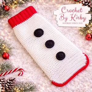 May include: A handmade crochet Christmas stocking in white with a red cuff and trim. Three black circular embellishments are sewn down the front. The stocking is decorated with Christmas ornaments and greenery.