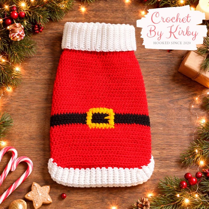 May include: A red and white crocheted Santa Claus outfit for a pet, featuring a black belt with a yellow buckle. The outfit has a white collar and trim. The text "Crochet By Kirby" is visible in the upper right corner.