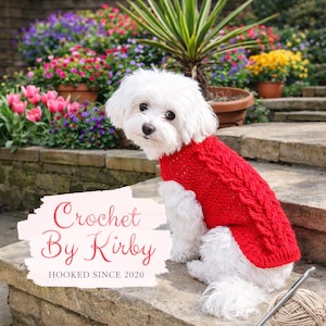Puede incluir: Un perro blanco pequeño con un suéter rojo de punto trenzado sentado en escalones de piedra. El suéter tiene un diseño texturizado. La imagen incluye el texto "Crochet By Kirby" y "HOOKED SINCE 2020". Flores y plantas coloridas en el fondo.