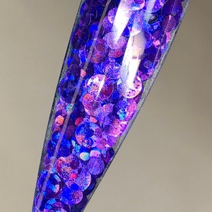May include: A close-up of a clear nail tip filled with iridescent purple and blue glitter.