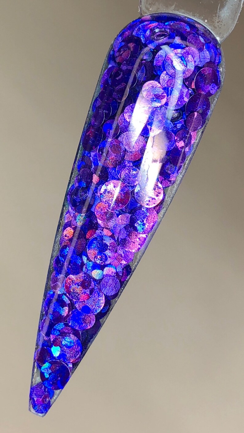 May include: A close-up of a clear nail tip filled with iridescent purple and blue glitter.