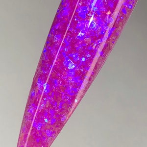 K&ouml;nnte beinhalten: Ein l&auml;nglicher, spitzer Nagel-Swatch zeigt einen schimmernden, magentafarbenen Nagellack. Der Lack ist mit irisierenden, facettenreichen Glitzerflocken durchzogen, die violette und blaue Farbt&ouml;ne reflektieren und einen schillernden Effekt erzeugen.