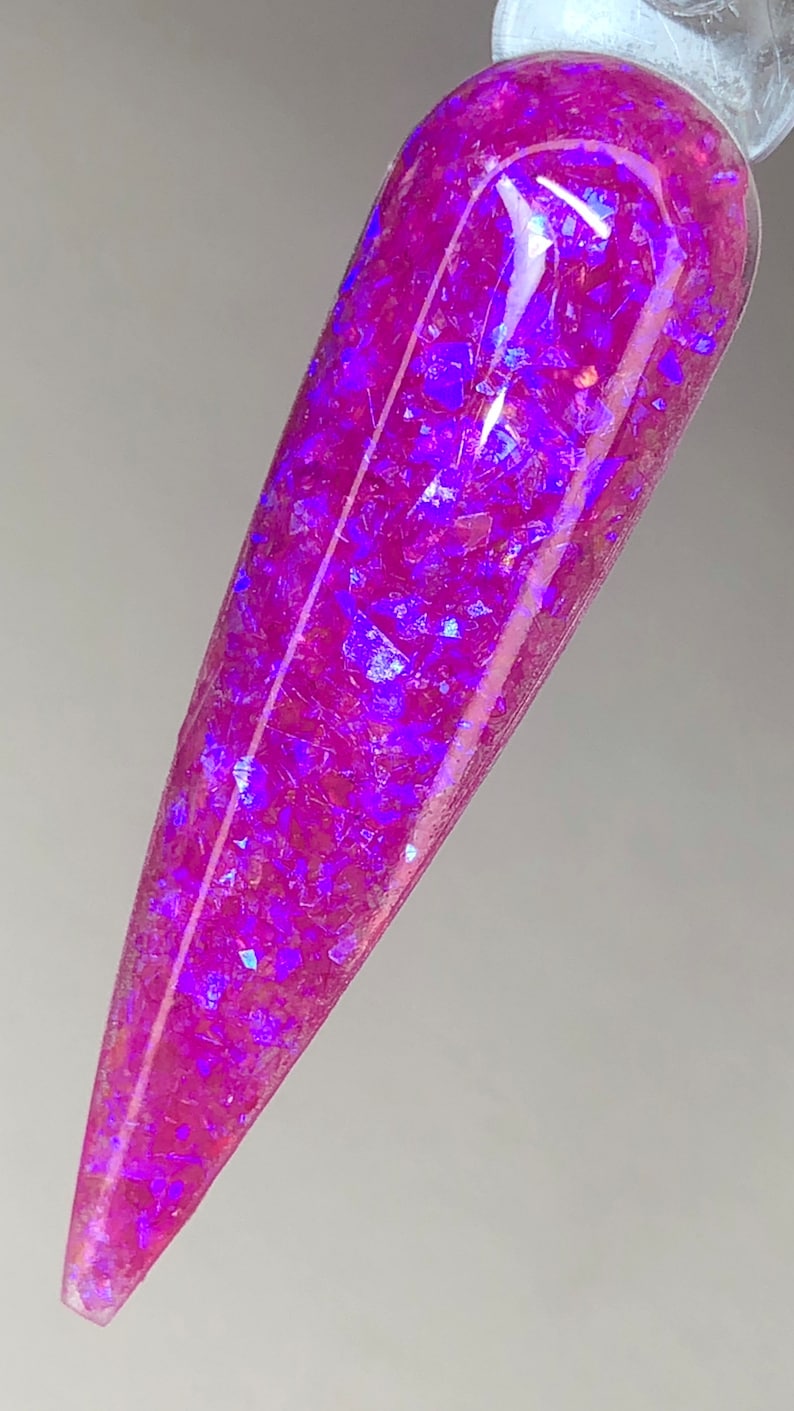 K&ouml;nnte beinhalten: Ein l&auml;nglicher, spitzer Nagel-Swatch zeigt einen schimmernden, magentafarbenen Nagellack. Der Lack ist mit irisierenden, facettenreichen Glitzerflocken durchzogen, die violette und blaue Farbt&ouml;ne reflektieren und einen schillernden Effekt erzeugen.