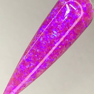 K&ouml;nnte beinhalten: Ein einzelner, langer, spitzer Nagel mit einem lebendigen, irisierenden lila Glitzerdesign. Der Nagel hat eine hellrosa Farbe mit gl&auml;nzendem Finish, und der Glitzer reflektiert das Licht und erzeugt einen funkelnden Effekt. Der Nagel ist vor einem schlichten wei&szlig;en Hintergrund platziert.