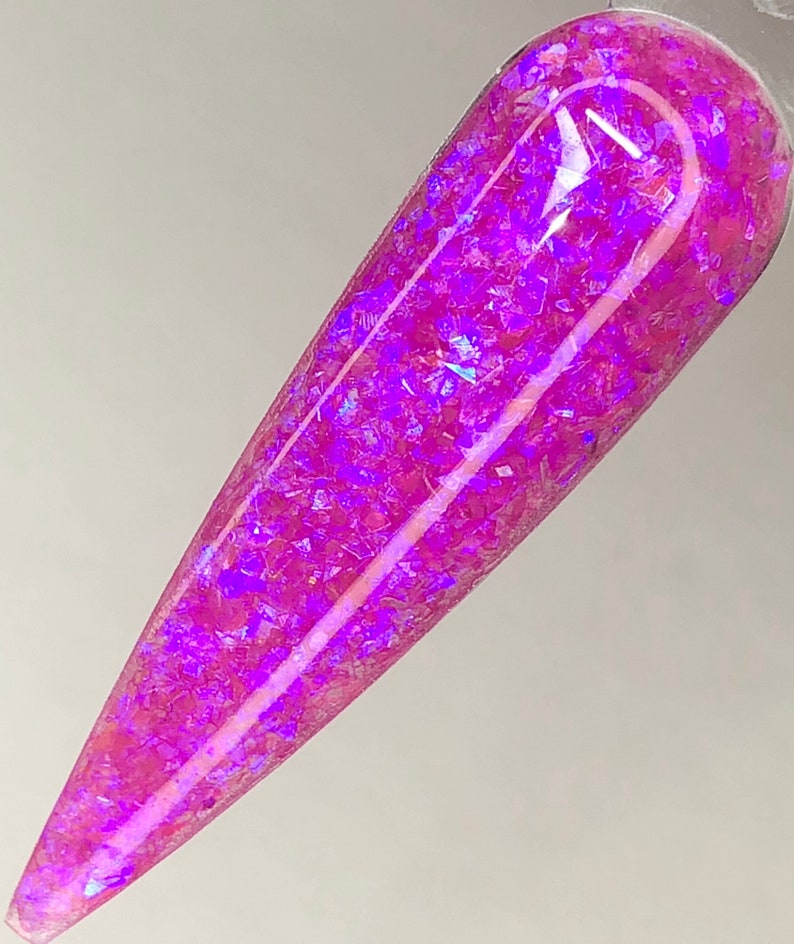 K&ouml;nnte beinhalten: Ein einzelner, langer, spitzer Nagel mit einem lebendigen, irisierenden lila Glitzerdesign. Der Nagel hat eine hellrosa Farbe mit gl&auml;nzendem Finish, und der Glitzer reflektiert das Licht und erzeugt einen funkelnden Effekt. Der Nagel ist vor einem schlichten wei&szlig;en Hintergrund platziert.