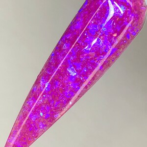 K&ouml;nnte beinhalten: Ein langer, spitzer Nagel mit leuchtend rosa Basis und eingebetteten irisierenden lila Glitzerflocken. Der Nagel wird vor einem hellen Hintergrund gehalten, der das gl&auml;nzende Finish und das Lichtspiel auf dem Glitzer zeigt.