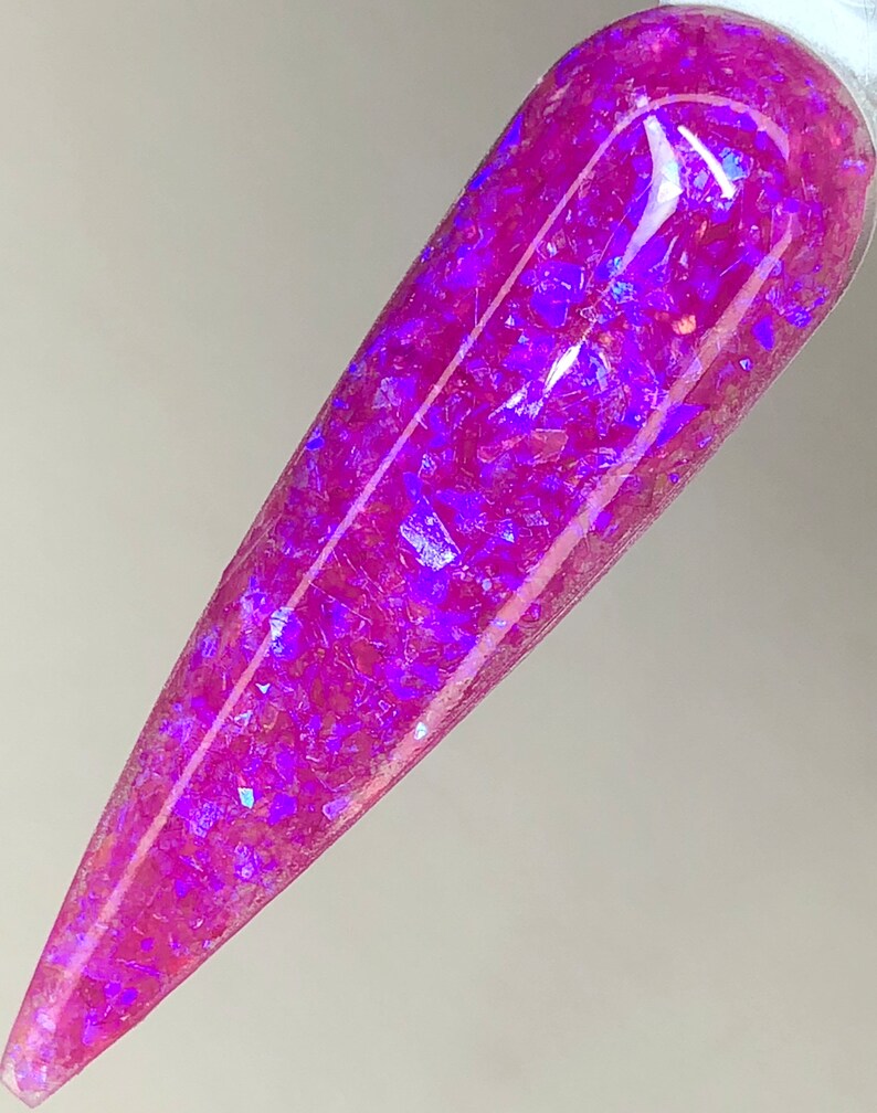 K&ouml;nnte beinhalten: Ein langer, spitzer Nagel mit leuchtend rosa Basis und eingebetteten irisierenden lila Glitzerflocken. Der Nagel wird vor einem hellen Hintergrund gehalten, der das gl&auml;nzende Finish und das Lichtspiel auf dem Glitzer zeigt.