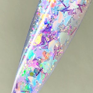 以下が含まれることがあります： 透明なネイルポリッシュボトルに、星、ハート、月など様々な形の虹色のグリッターが入っています。グリッターはピンク、ブルー、パープルの色合いです。