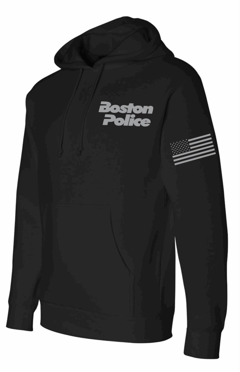 Puede incluir: Sudadera con capucha negra con las palabras "Boston Police" en letras plateadas en la parte delantera y una bandera estadounidense plateada en la manga derecha.