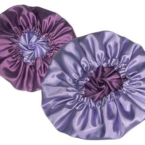 Op de afbeelding: Twee satijnen slaapmutsen, &eacute;&eacute;n paars en &eacute;&eacute;n lavendel, met een contrasterende voering. De slaapmutsen zijn ontworpen om het haar te beschermen tijdens het slapen.