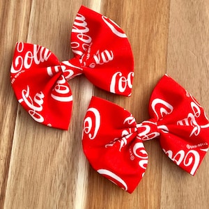 May include: Two red fabric hair bows with a white Coca-Cola logo print.