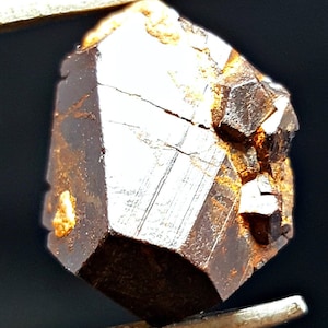 Puede incluir: Un cristal metálico marrón oscuro con forma geométrica. El cristal tiene una superficie rugosa y texturizada y parece estar parcialmente cubierto por una capa de óxido.