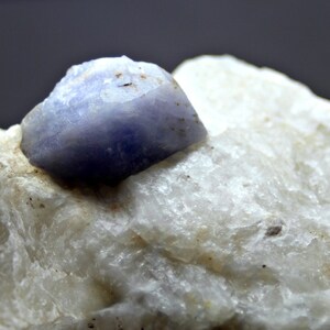 May include: A blue sapphire crystal on a white rock. The sapphire is a deep blue color and has a smooth, polished surface. The rock is rough and textured.