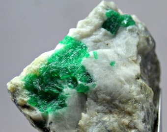 May include: A rough, white rock with bright green emerald crystals embedded in it. The emerald crystals are clustered together in a few areas of the rock.