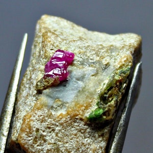 Puede incluir: Una pequeña roca marrón rugosa con un cristal de rubí rosa brillante incrustado. El rubí está rodeado de cristales más pequeños, verdes y dorados.