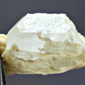 Puede incluir: Un cristal de cuarzo claro y facetado con una base beige lechosa. El cristal está sujeto por unas pinzas, mostrando su forma natural y claridad. El cuarzo tiene un aspecto rugoso y texturizado.