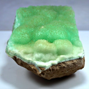 May include: A green and white mineral specimen with a rough, textured surface. The mineral is attached to a brown rock.