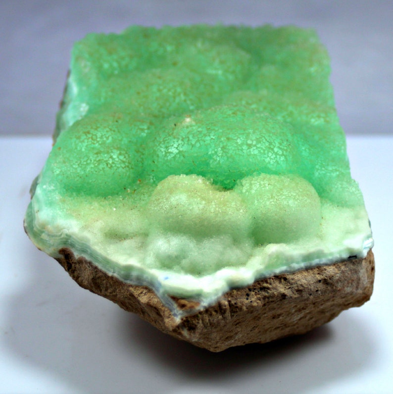 May include: A green and white mineral specimen with a rough, textured surface. The mineral is attached to a brown rock.