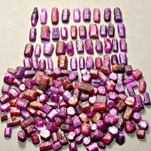 May include: A collection of raw ruby gemstones in various sizes and shapes. The gemstones range in color from deep red to purplish-pink, with some showing a mix of hues. The rough-cut stones are arranged on a gray surface.