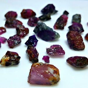Puede incluir: Una colección de piedras preciosas en bruto de color rosa y morado. Las piedras son de varios tamaños y formas, y algunas tienen un color oscuro, casi negro.
