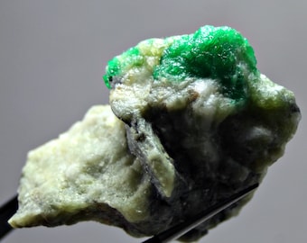 May include: A rough, green emerald crystal embedded in a white and gray rock matrix. The emerald has a bright, vibrant green color and is partially covered by the rock.