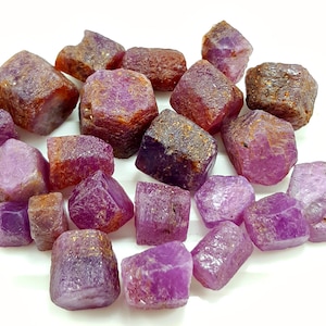 以下が含まれることがあります： ピンクとパープルのルビーの原石のコレクション。石は様々なサイズと形があり、一部はわずかに金属光沢があります。