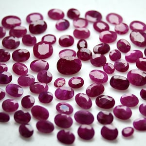 Pode incluir: Uma coleção de rubis vermelhos escuros de corte oval, de vários tamanhos, espalhados sobre uma superfície branca. As pedras preciosas têm uma superfície polida e refletora, mostrando suas facetas internas. Apresentação atraente.