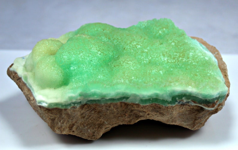 May include: A green and white mineral specimen with a rough, textured surface. The mineral is attached to a brown rock.
