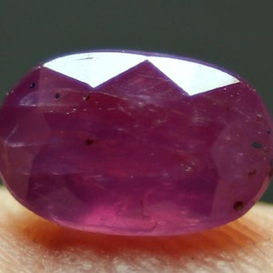 Könnte beinhalten: Ein facettierter, ovalförmiger Rubin-Edelstein. Der Edelstein ist ein tiefes, sattes Rot mit einer polierten Oberfläche, die das Licht reflektiert. Der Schliff zeigt mehrere Facetten, die seinen Glanz verstärken. Kleine Einschlüsse sind im Stein sichtbar.