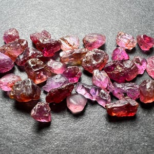 Puede incluir: Una colección de gemas en bruto, sin cortar, en tonos rosa, púrpura y marrón rojizo. Las piedras varían en tamaño y forma, con una textura rugosa y natural. Las gemas están dispersas sobre una superficie gris oscuro.