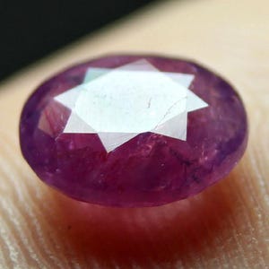 Può includere: Primo piano di una gemma di rubino ovale e sfaccettata. La gemma è di un colore magenta intenso, con un motivo a stella al centro. Le sfaccettature della pietra catturano la luce.