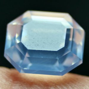 May include: A light blue, octagonal gemstone with multiple facets. The gem is transparent, allowing light to pass through and reflect off the cut surfaces. The gemstone is set against a dark background.