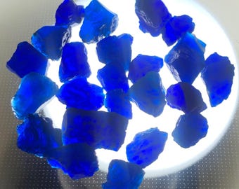Puede incluir: Una colección de pequeños trozos de vidrio azul cobalto, irregulares y translúcidos, con bordes dentados y acabado brillante. Las piezas están esparcidas sobre una fuente de luz blanca circular que ilumina su color azul intenso.