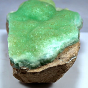May include: A green and white mineral specimen with a layered, crystalline structure. The mineral is attached to a brown rock base.