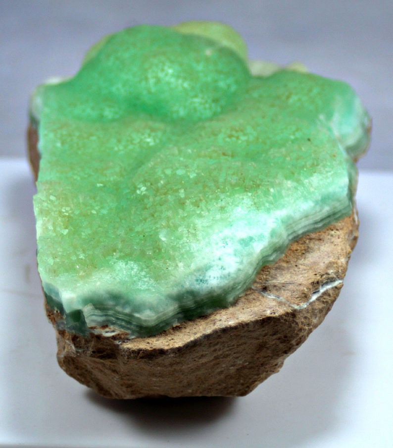 May include: A green and white mineral specimen with a layered, crystalline structure. The mineral is attached to a brown rock base.