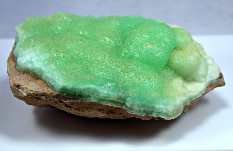 May include: A green and white mineral specimen with a layered, bubbly texture. The mineral is attached to a brown rock.