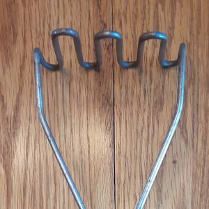 May include: A vintage potato masher with a green handle and a white stripe. The metal mashing head has a wavy design. The kitchen utensil is set against a wooden background.