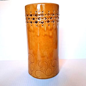 May include: A tall, cylindrical vase with a glossy, amber-colored finish. The upper portion features a series of decorative cut-out floral patterns. The base is adorned with a swirling, embossed design. The vase is approximately 12 inches tall.