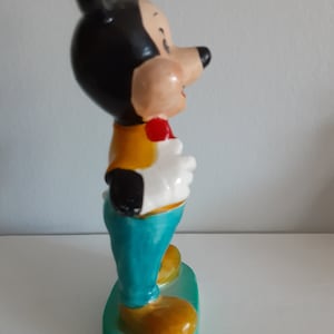 Op de afbeelding: Een keramieken beeldje van Mickey Mouse, met een zwarte kop en oren, een beige gezicht en een rode vlinderdas. Hij draagt een geel vest, witte handschoenen en een turquoise broek. Het beeldje staat op een groene basis met gele schoenen.