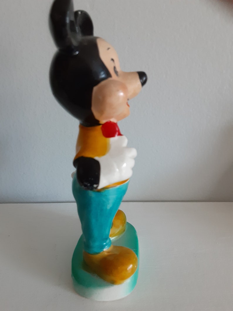 Op de afbeelding: Een keramieken beeldje van Mickey Mouse, met een zwarte kop en oren, een beige gezicht en een rode vlinderdas. Hij draagt een geel vest, witte handschoenen en een turquoise broek. Het beeldje staat op een groene basis met gele schoenen.