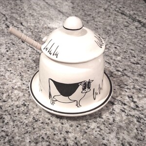 May include: A white ceramic sugar bowl with a black and white cow illustration. The bowl has a lid and sits on a matching saucer. A small wooden spoon rests on the side. The bowl is decorated with cursive script.